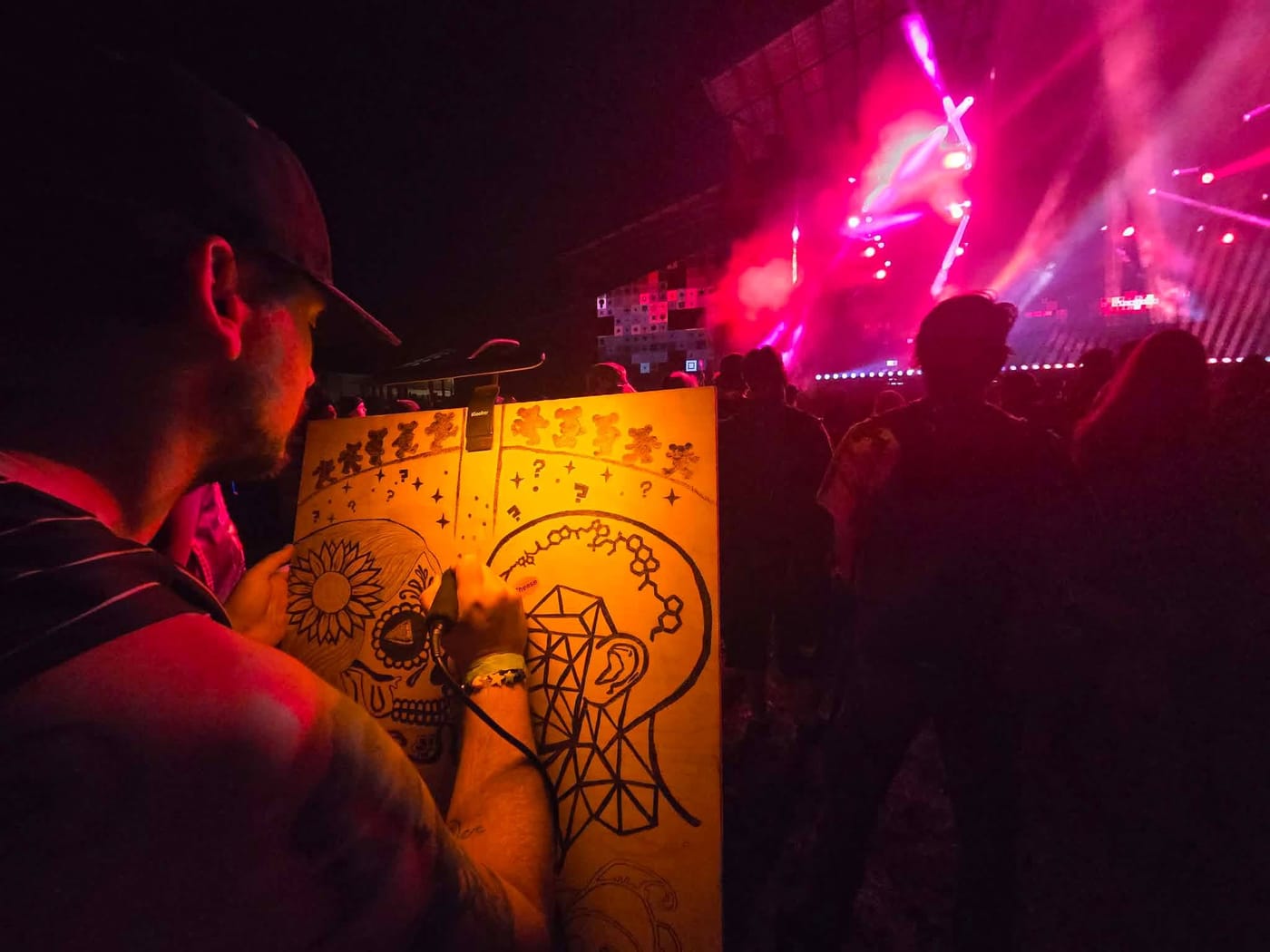 The artist burning a skull and geometric head design into a wood panel in front of a pink-lit festival stage