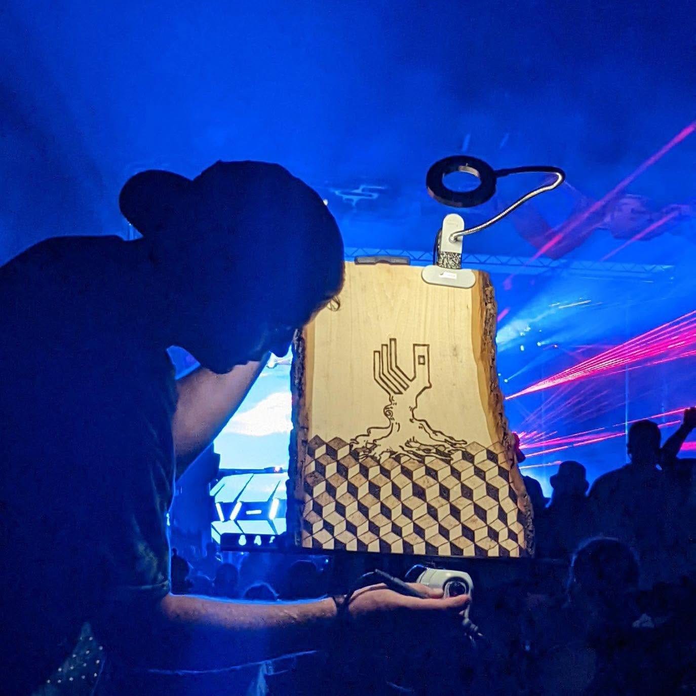 The artist burning a tree and cube pattern into a raw edge wood slab in front of a blue-lit stage
