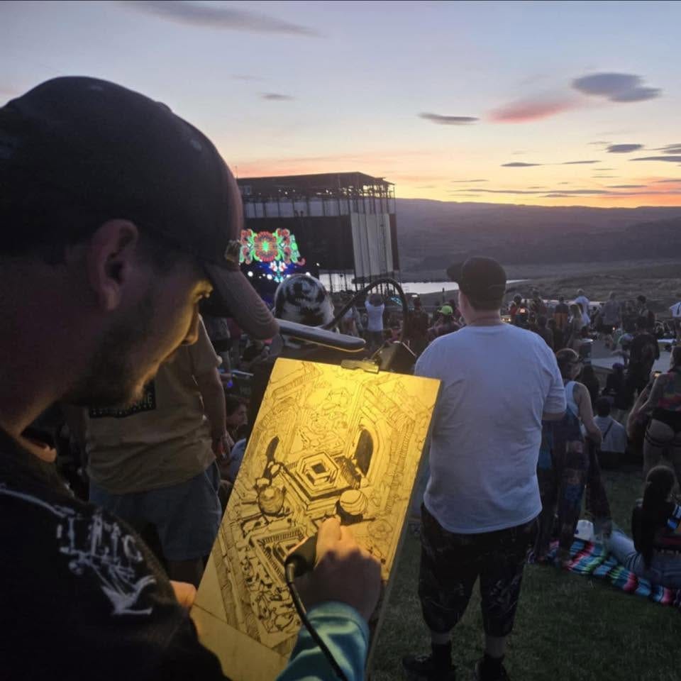 The artist burning an isometric design into a wood panel at sunset in front of a festival stage